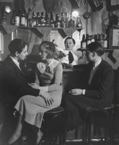 Image depicts women defying rigid social structures and rules about acceptable women's attire in 1932. Three women are seated at the bar at Le Monocle in Paris in 1932. A woman bartender is in the background, wearing a shirt and tie and short hair. Two of the women are dressed in masculine attire, with suits and ties and slicked back short hair. One of the women is in a dress or a sweater and long skirt, with shoulder length hair in soft curls and a hat. These iconic black-and-white gelatin silver prints document the vibrant queer culture of Paris at Le Monocle lesbian bar in Montmartre. Noticeable is the impeccable tailoring, crisp lapels, and clean architecture of the formal attire.