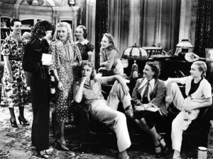 Women characters, played by actresses Eve Arden, Katharine Hepburn, Ginger Rogers, and the ensemble, are seated in the boarding house lobby in the film Stage Door, illustrating acceptable attire fitted to one's personality. Some women are wearing skirts and dresses and others are wearing trousers, masculine suits and ties, all looking relaxed and comfortable in their own skin. This is a study in casual, communal symmetry and the unhurried liberty of women living on their own terms.